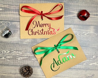 May include: Two kraft paper gift envelopes with red and green satin ribbons. One envelope has "Merry Christmas" in red foil, the other has "Adam" in green foil. A red jingle bell and pine cone are nearby.