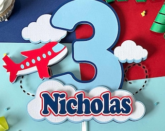 May include: A blue and red airplane cake topper with the number 3, clouds, and the name "Nicholas". The airplane is red with white and blue accents. The number 3 is light blue with a dark blue outline. The name is in a white cloud.