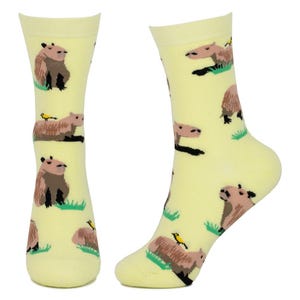 以下が含まれることがあります： 薄い黄色の靴下のペア。茶色のカピバラと黄色の鳥が緑の草の上に繰り返しのパターンで描かれています。靴下は膝丈で、柔らかい素材で作られています。