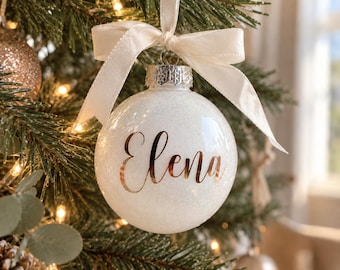May include: A spherical white glitter Christmas ornament featuring the name Elena in metallic rose gold script lettering. The ornament is topped with a silver metal cap and a tied cream-colored satin ribbon for hanging on a tree branch.