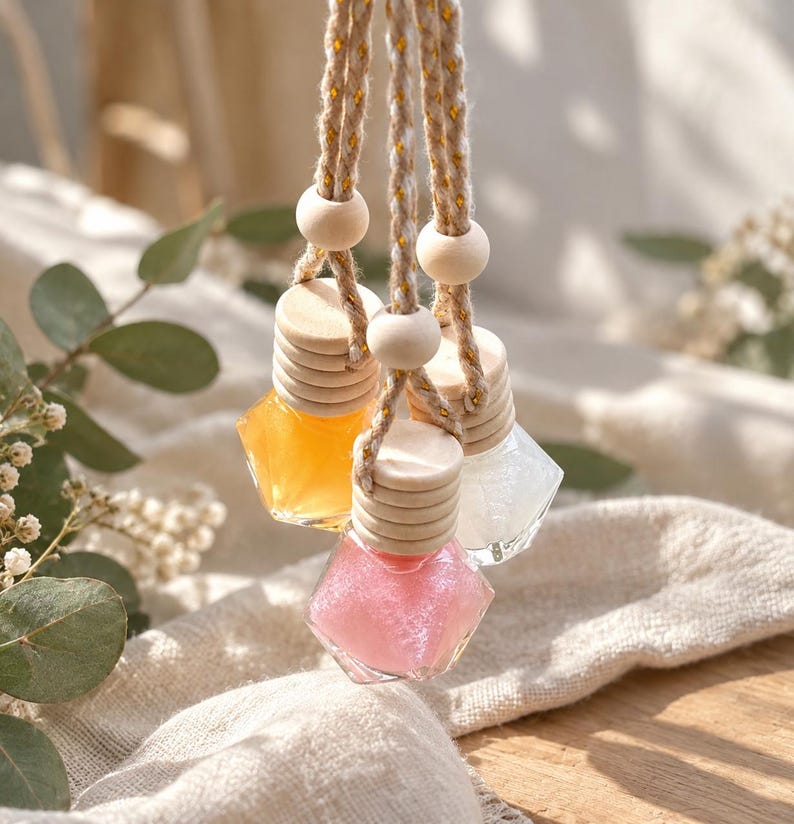 May include: Three small, diamond-shaped glass bottles filled with orange, pink, and white liquid. Each bottle has a light-colored wooden cap and is suspended by a braided beige cord featuring metallic gold threads and round wooden beads.