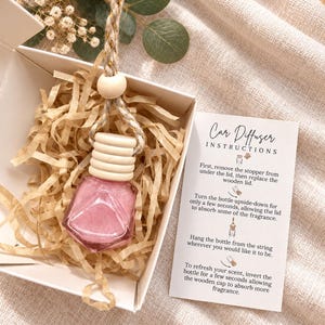 May include: A pink diamond-shaped glass car diffuser bottle with a ribbed wooden cap and a braided hanging cord, nestled in a white gift box with paper filler. An instruction card titled Car Diffuser Instructions is included.