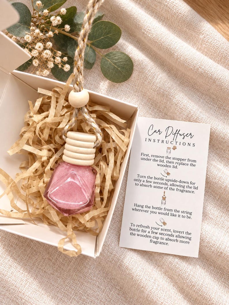 May include: A pink diamond-shaped glass car diffuser bottle with a ribbed wooden cap and a braided hanging cord, nestled in a white gift box with paper filler. An instruction card titled Car Diffuser Instructions is included.