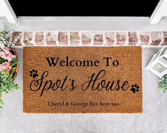 May include: A brown coir doormat with the text "Welcome To Spot's House" in black cursive lettering. Two black paw prints are on either side of the text. The text "Cheryl & George live here too" is printed in black below the main text.