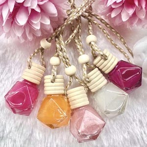 May include: Five small, diamond-shaped glass bottles filled with colourful, shimmering liquids. Each bottle features a ribbed wooden cap and is suspended by a braided tan cord with a small wooden bead. The colours include magenta, orange, light pink, and clear.