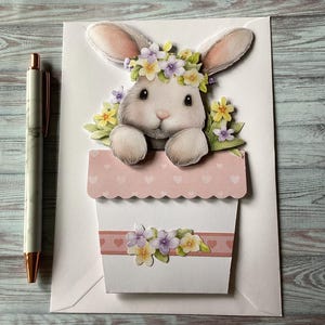 Peut inclure: Une carte de Pâques faite à la main avec une illustration 3D d'un lapin blanc portant une couronne de fleurs, sortant d'un pot de fleurs rose et blanc. La carte est décorée de fleurs pastel et se trouve sur une enveloppe blanche, à côté d'un stylo.