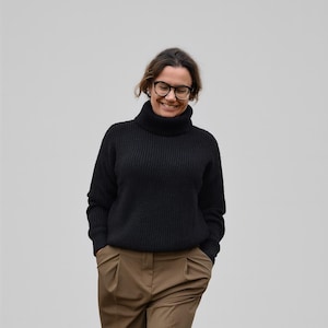 Puede incluir: Un jersey negro de cuello alto y pantalones marrones con pliegues. El jersey tiene una textura acanalada. Los pantalones tienen cintura alta y un corte holgado. La persona lleva gafas y sonríe.