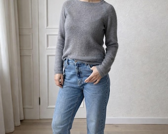 May include: A long-sleeved, crew-neck sweater in a soft, heathered gray knit fabric paired with light-wash blue denim jeans. The jeans feature a high-waisted fit, a button and zipper fly closure, and are cuffed at the bottom hem above the ankles.