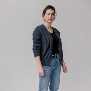 May include: A woman wearing a dark gray cardigan over a black top and light blue jeans. She is also wearing a black belt and black boots. The cardigan has a V-neck and button closure. The jeans are straight-legged.