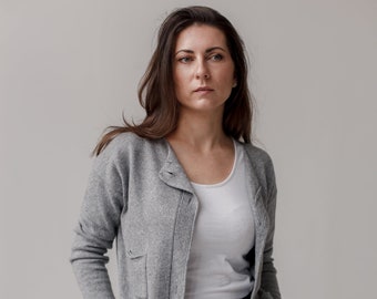 May include: A light gray knit cardigan with a round neckline, long sleeves, and two small front pockets. The cardigan is worn over a plain white scoop neck tank top and paired with solid black trousers that feature a button and zipper closure.