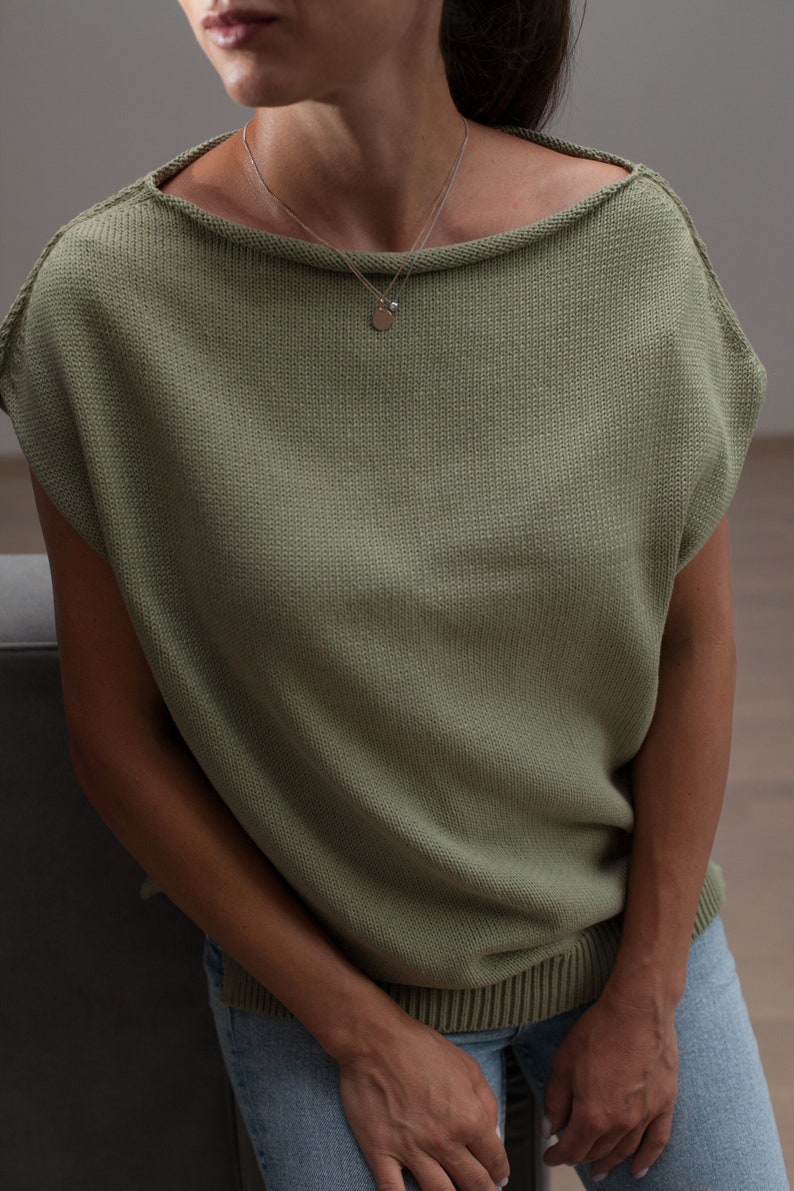 Puede incluir: Una mujer lleva una blusa de punto verde claro con cuello de barco y mangas cortas. Lleva un collar plateado con un peque&ntilde;o colgante.