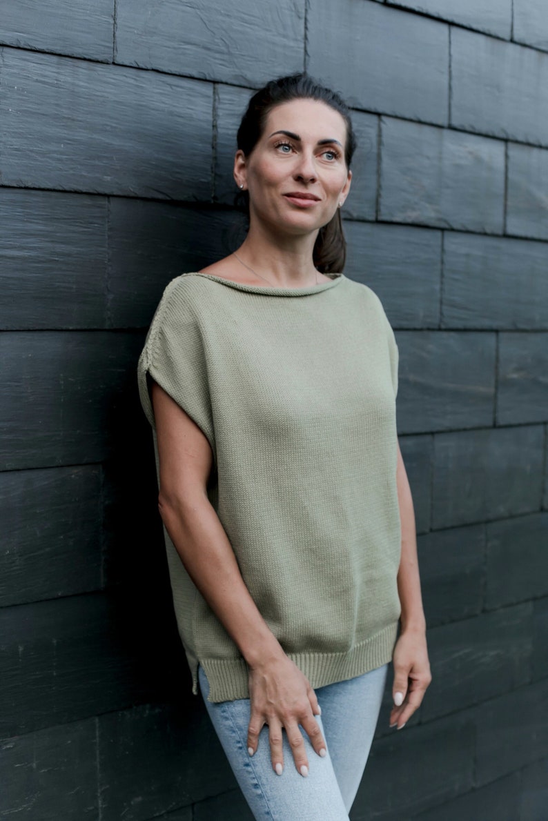 Puede incluir: Una mujer con un top de punto verde claro de manga corta y pantalones vaqueros azules. Est&aacute; de pie frente a una pared de ladrillo negro.