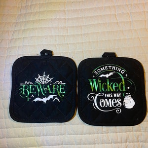 May include: Two black quilted pot holders with white and green text. One pot holder says "Beware" with a spider web and bats. The other pot holder says "Something Wicked This Way Comes" with a cauldron.