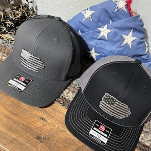 May include: Two adjustable Richardson 112 trucker hats. One is dark gray with a black mesh back, the other is black with a gray mesh back. Both have a distressed American flag patch. A blue flag with white stars is in the background.