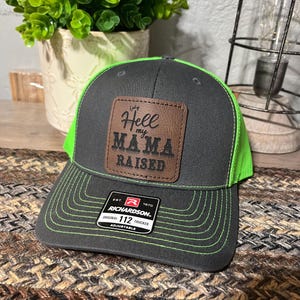 May include: A gray and neon green trucker hat with the text "Hell my Mama Raised" on a brown leather patch. The hat is a Richardson 112 Trucker style and is adjustable.