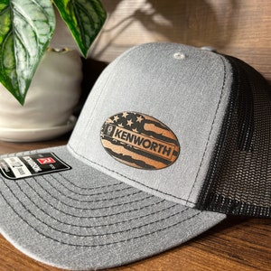May include: A grey trucker hat with a black mesh back and a flat brim. Centered on the front is an oval-shaped brown leather patch featuring a laser-engraved American flag design and the text KENWORTH. A black product tag is attached to the brim.