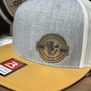 May include: Gray and mustard yellow trucker hat with a mesh back. The hat features a leather patch with a skull and the text "FUCK AROUND AND FIND OUT". The brim is mustard yellow, and the front panel is gray.
