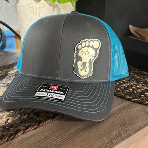May include: A charcoal gray trucker hat with bright blue mesh back panels and matching blue stitching on the brim. A tan patch on the front features a footprint silhouette containing a mountain landscape and a Bigfoot figure. A black Richardson 112 label is attached to the brim.