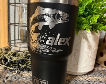 May include: Black and silver insulated tumbler with a fish graphic and the name "Alex". The tumbler has a silver rim and a black base, with the brand name "YETI" displayed prominently. A decorative item for fishing enthusiasts.