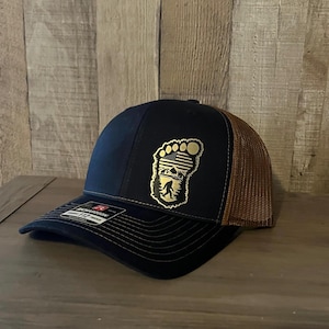 May include: A navy blue baseball cap with a brown mesh back. The cap features a gold-colored Bigfoot footprint design with an American flag and mountain scene. The brim is curved, and the cap has a Richardson 112 tag.
