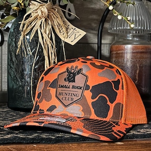 May include: Orange and black camouflage trucker hat with a mesh back. The front of the hat features a patch with a deer silhouette and the text "SMALL BUCK HUNTING CLUB".