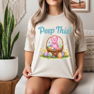May include: Beige t-shirt with the text "Peep This!" in light blue. The graphic features a pink bunny in a basket with Easter eggs, flowers, and chicks. The shirt is a casual, short-sleeved style.