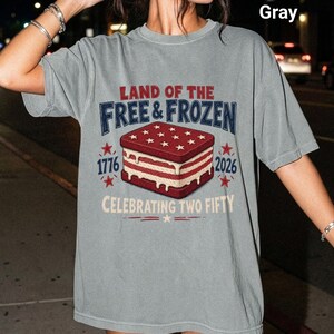 May include: A gray short-sleeve t-shirt featuring a graphic of an ice cream sandwich with red and white layers. The text reads LAND OF THE FREE & FROZEN, 1776, 2026, and CELEBRATING TWO FIFTY, accompanied by red star icons and decorative lines.