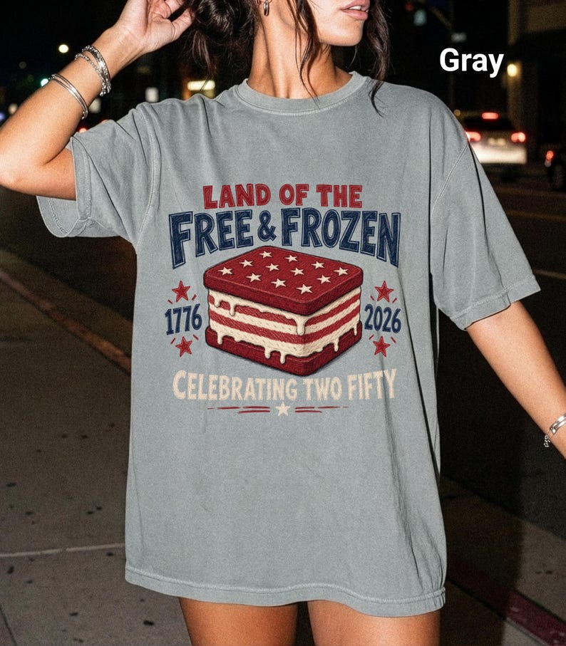 May include: A gray short-sleeve t-shirt featuring a graphic of an ice cream sandwich with red and white layers. The text reads LAND OF THE FREE & FROZEN, 1776, 2026, and CELEBRATING TWO FIFTY, accompanied by red star icons and decorative lines.