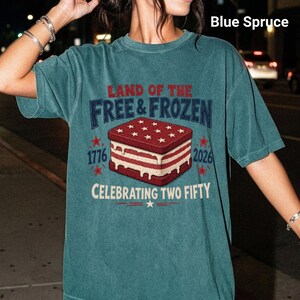 May include: A blue spruce short-sleeve t-shirt featuring a graphic of a red, white, and blue ice cream sandwich. The text reads: LAND OF THE FREE & FROZEN, 1776, 2026, and CELEBRATING TWO FIFTY. The design includes stars and red accents.