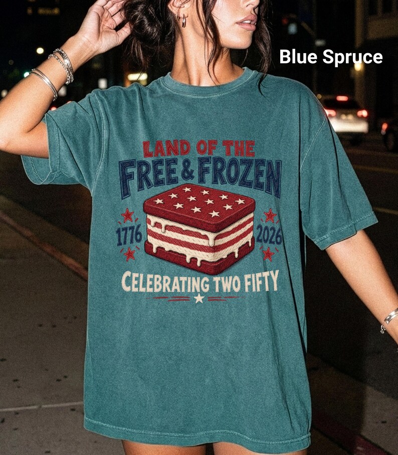 May include: A blue spruce short-sleeve t-shirt featuring a graphic of a red, white, and blue ice cream sandwich. The text reads: LAND OF THE FREE & FROZEN, 1776, 2026, and CELEBRATING TWO FIFTY. The design includes stars and red accents.