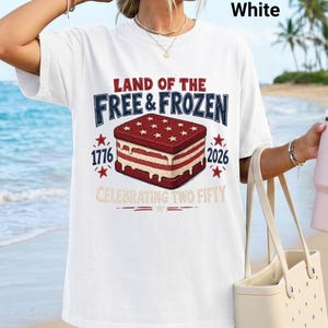 May include: A white short-sleeve t-shirt featuring a graphic of an ice cream sandwich with red and white stripes and stars. The text reads LAND OF THE FREE & FROZEN, 1776, 2026, and CELEBRATING TWO FIFTY. The design includes red stars and decorative lines.