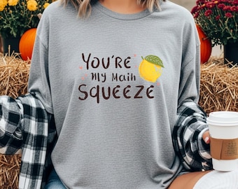 Könnte beinhalten: Ein graues Langarmshirt mit dem Text "You're my Main Squeeze" und einer Zitronenillustration. Die Person trägt ein schwarz-weiß kariertes Hemd und hält eine Kaffeetasse. Kürbisse und Blumen sind im Hintergrund.