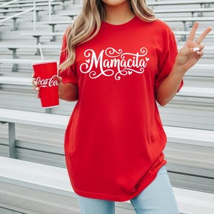 Pode incluir: Camiseta vermelha com a palavra "Mamcita" em escrita branca. A camisa é combinada com jeans azul claro. Uma xícara vermelha de Coca-Cola com um canudo branco é segurada na mão esquerda.