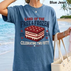 May include: A blue jean colored t-shirt featuring a graphic of a red, white, and blue ice cream sandwich. The text reads LAND OF THE FREE & FROZEN, 1776, 2026, and CELEBRATING TWO FIFTY. The design includes stars and melting ice cream details.