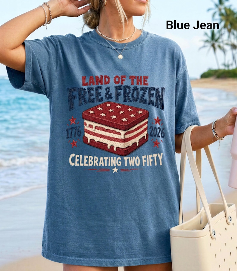 May include: A blue jean colored t-shirt featuring a graphic of a red, white, and blue ice cream sandwich. The text reads LAND OF THE FREE & FROZEN, 1776, 2026, and CELEBRATING TWO FIFTY. The design includes stars and melting ice cream details.