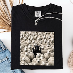 Puede incluir: Una camiseta negra con un estampado gráfico de un gran rebaño de ovejas blancas rodeando a una oveja negra en el centro. La oveja negra aparece con los dos dedos corazón levantados. La camiseta tiene una etiqueta de la marca Comfort Colors.