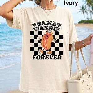 May include: Ivory-coloured t-shirt featuring the text "Same Weenie Forever" and a cartoon hot dog graphic. The graphic shows a hot dog with a hat, set against a black and white checkered background. The shirt is a casual, oversized style.