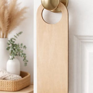 Könnte beinhalten: Ein leerer Türhänger aus Holz mit einem kreisförmigen Ausschnitt oben, der an einem Messing-Türknauf hängt. Der rechteckige Türhänger hat abgerundete Ecken. Der Hintergrund zeigt eine weiße Tür und eine dekorative Innenausstattung.