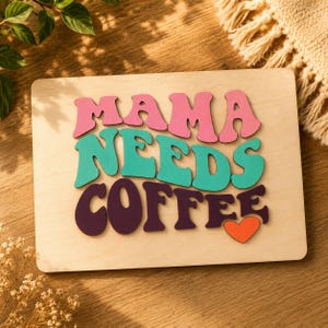 Könnte beinhalten: Holzschild mit dem Schriftzug "MAMA NEEDS COFFEE" in rosa, türkis und lila Buchstaben, mit einem orangefarbenen Herz. Das Schild befindet sich auf einem hellen Holzgrund.