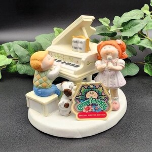 May include: A white porcelain figurine of two Cabbage Patch Kids playing music. The girl is holding a microphone and the boy is playing a white piano. A small brown and white dog sits next to the piano. The figurine has a Cabbage Patch Kids label that reads "Special Limited Edition".
