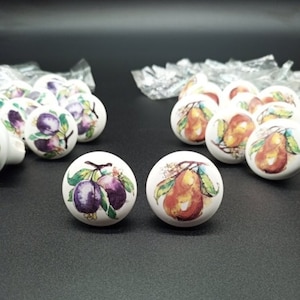 May include: A set of 12 white ceramic drawer knobs with a fruit design. The knobs feature pears and plums with green leaves. The knobs are approximately 1 inch in diameter.