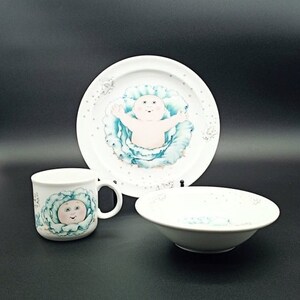 May include: A white ceramic set of a plate, bowl, and mug featuring a Cabbage Patch Kid doll in a cabbage. The plate and bowl have a white background with a blue cabbage and a white doll. The mug has a white background with a blue cabbage and a white doll's face.