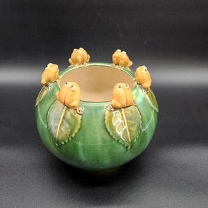 May include: A green ceramic bowl with a round shape, featuring frog figurines around the rim. The frogs are a light orange color and sit atop green leaves. The bowl has a light brown interior.