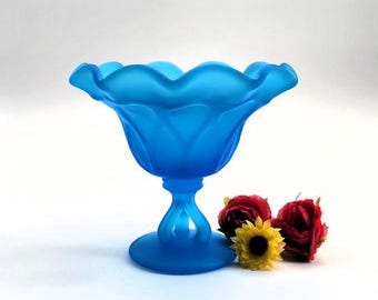 May include: A frosted blue glass bowl with a scalloped rim and a pedestal base. The bowl is sitting on a white surface with red and yellow flowers.