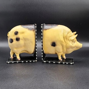 May include: A pair of pig-shaped bookends. The bookends are painted yellow with black spots and a dark brown top. They stand on a black base with a black and white striped edge. The pig bookends are a decorative item.