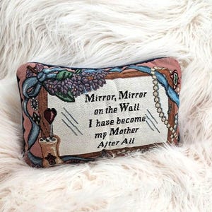May include: A rectangular tapestry pillow with the text "Mirror, Mirror on the Wall I have become my Mother After All." The pillow has a pink and brown border with floral and ribbon designs. A string of pearls is on the right side.