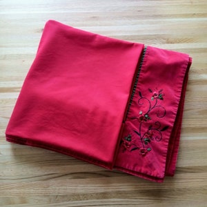 May include: Red tablecloth with a floral embroidery design in black and green thread. The embroidery is on one edge of the tablecloth.