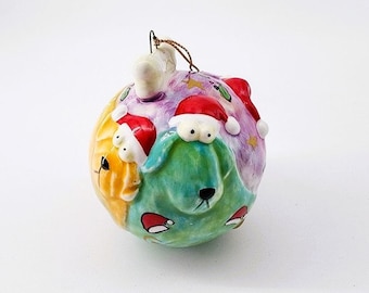 May include: A ceramic Christmas ornament shaped like a ball with three dogs wearing Santa hats. The dogs are painted in shades of green, yellow, and purple.