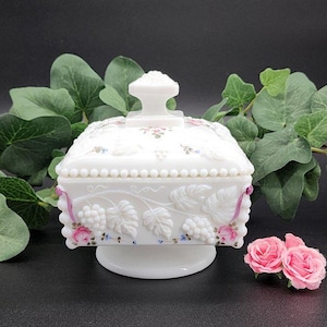 May include: A white, square, lidded dish with a pedestal base. The dish features embossed grape and leaf designs with pink and blue accents. The lid has a decorative handle and floral details. A pair of pink roses sits nearby.