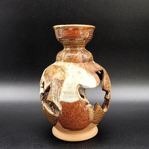 May include: A handcrafted ceramic vase with a unique, openwork design. The vase features a combination of brown, tan, and white glazes, creating a textured appearance. The vase has a round body with cut-out shapes and a flared top, standing on a small, circular base.